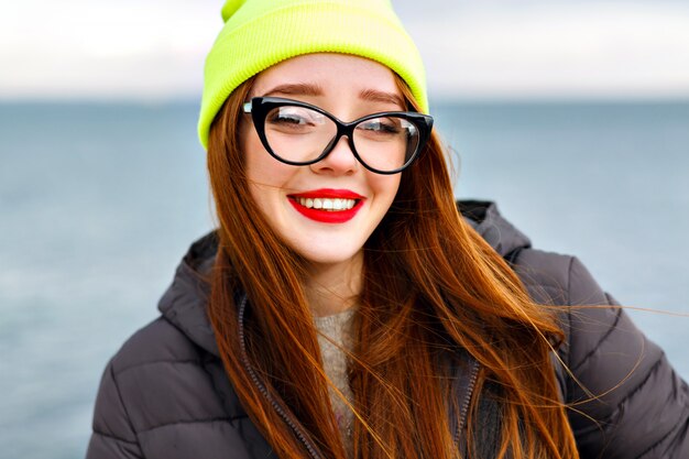 Bliska portret uroczej uśmiechniętej wesołej kobiety z rudymi włosami, spędzającej niesamowity czas na plaży, zimowy czas, młody podróżnik, jasny makijaż, neonowa czapka, ciepła kurtka, okulary hipster.