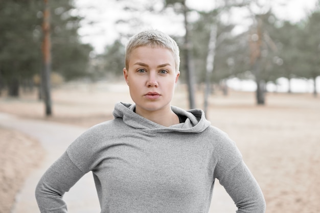 Bliska Strzał Piękna Sportowa Kobieta Ubrana W Bluzę Z Kapturem Podczas Porannego Spaceru Na świeżym Powietrzu, Pozowanie Na Tle Niewyraźne Sosny. Koncepcja Ludzie, Aktywny Tryb życia, Zdrowie, Sport I Fitness