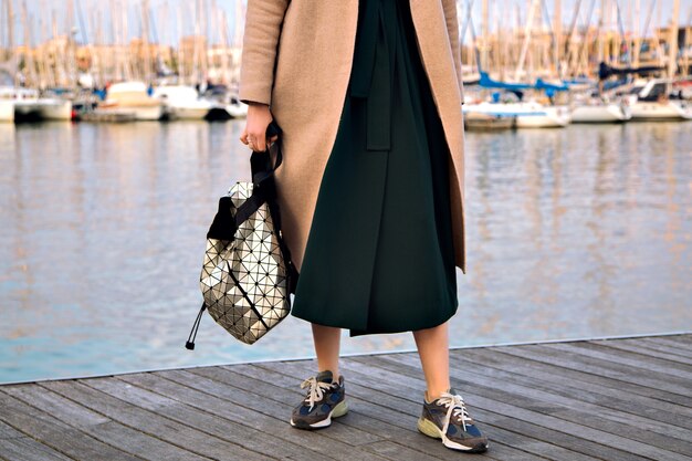 Bliska szczegóły mody modnej kobiety noszącej elegancką sukienkę, nowoczesne modne trampki i plecak z eleganckim kaszmirowym płaszczem, pozuje na nadmorskiej promenadzie, w połowie sezonu, w delikatnych pastelowych kolorach.