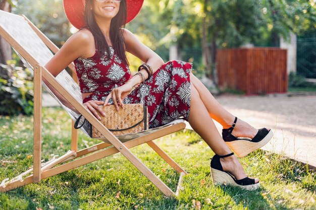 Bliska szczegóły nogi w sandałach na koturnie, obuwie, stylowa piękna kobieta siedząca na leżaku w stroju w stylu tropikalnym, trend w modzie letniej, torebka ze słomy, akcesoria, wakacje