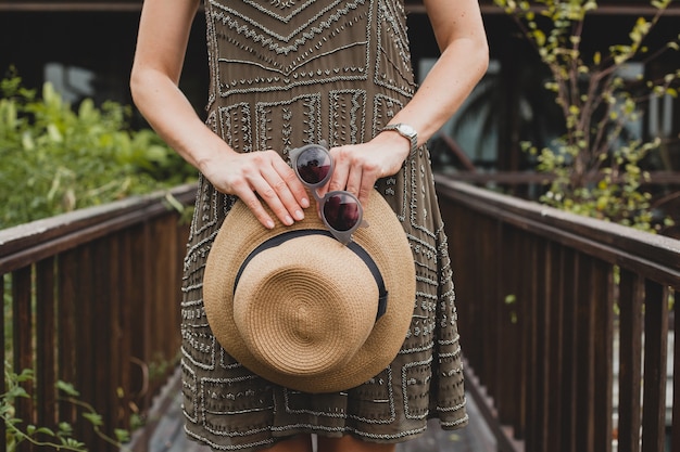 Bliska szczegóły ręce trzymając słomkowy kapelusz i okulary przeciwsłoneczne, stylowe akcesoria, młoda atrakcyjna kobieta w eleganckiej sukience, letni styl, trend w modzie, wakacje, pozowanie na tropikalnej willi, uśmiechnięta, szczęśliwa