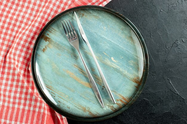 Bliska widok z góry stalowy widelec i nóż obiadowy na okrągłym talerzu czerwony i biały obrus w kratkę na czarnym stole