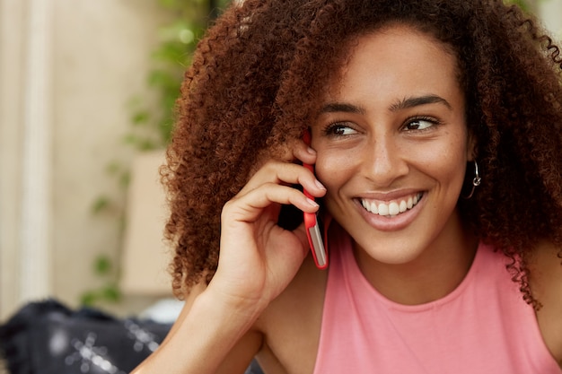 Bliskie Ujęcie Przyjemnie Wyglądającej Młodej Wesołej Kobiety Z Fryzurą Afro, Cieszącej Się, że Słyszy Głos Chłopaka Przez Nowoczesny Telefon Komórkowy, Nie Widziano Jej Od Dawna, Bardzo Tęsknimy Za Sobą