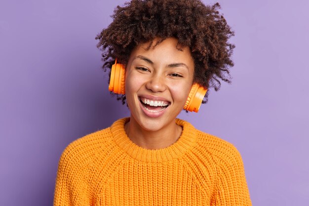 Bliskie ujęcie uradowanej Afro American kobieta uśmiecha się szeroko i ma kręcone włosy, nosi stereofoniczne słuchawki bezprzewodowe ubrane w dzianinowy pomarańczowy sweter