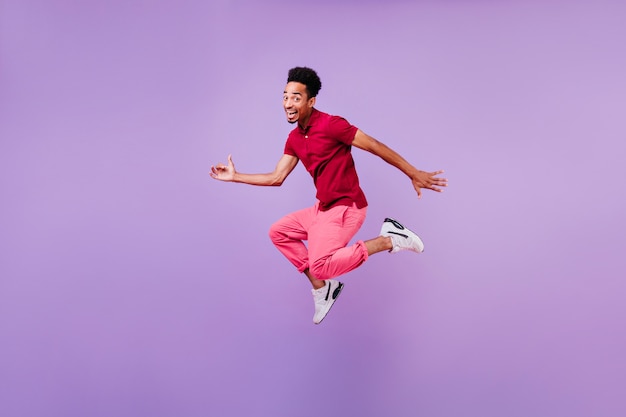 Blithesome czarny brunetka mężczyzna tańczy z radosnym uśmiechem. Wewnątrz zdjęcie inspirowanego faceta w czerwonych spodniach i białych butach skaczących.