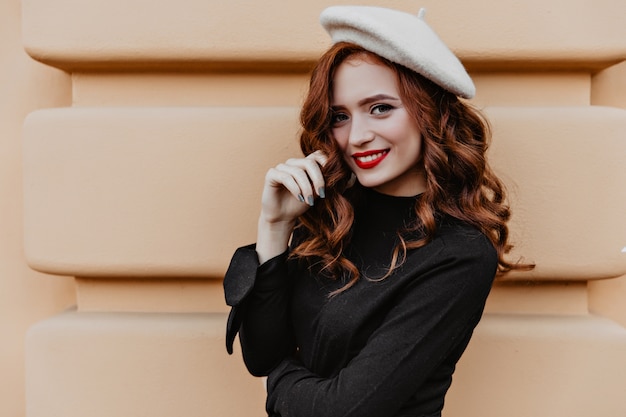 Bezpłatne zdjęcie blithesome kaukaski dziewczyna w czarnej bluzce pozuje na zewnątrz. oszałamiająca młoda kobieta w brązowym berecie stojąca blisko ściany z uśmiechem.