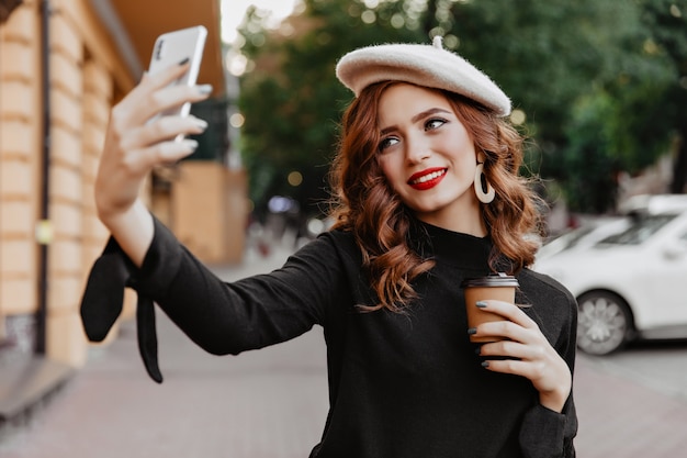 Blithesome rudowłosa dziewczyna trzyma filiżankę kawy i pozowanie na zewnątrz. Beztroska francuska młoda dama robi selfie na ulicy.