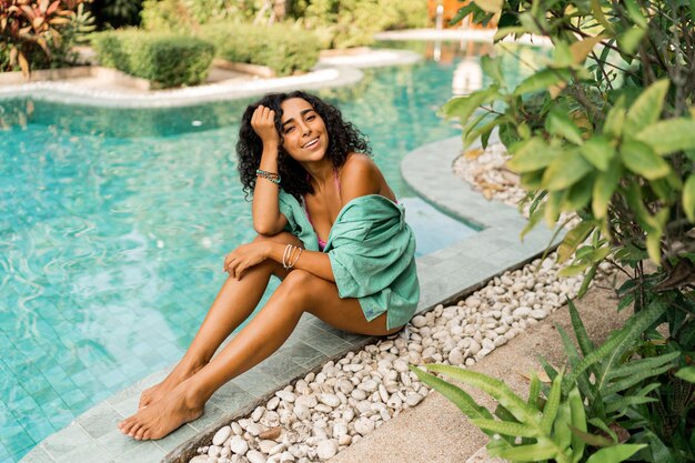 Błoga kobieta z falującymi włosami w letnim stroju siedząca przy basenie Nastrój wakacji i podróży Jasne kolory Modne dodatki