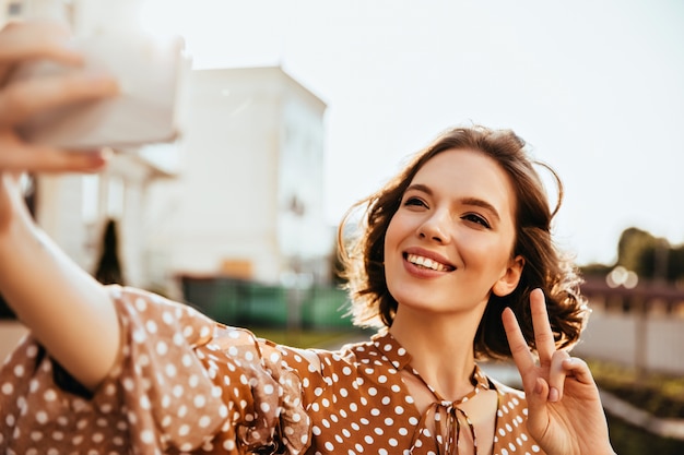 Błoga krótkowłosa kobieta w brązowych ubraniach wyrażająca szczęście. Jocund pani trzyma telefon i robi selfie.