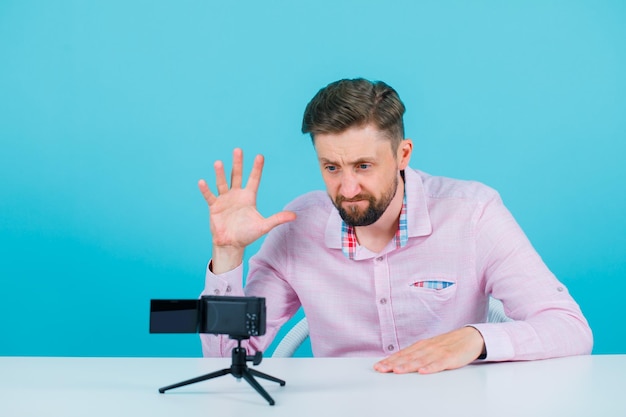 Blogger mężczyzna pozuje do swojej mini kamery, pokazując gest pazurów na niebieskim tle