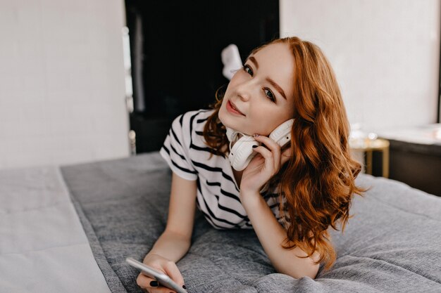 Błogi rudowłosa dziewczyna leżąc na łóżku z telefonem. Zainteresowana kaukaski kobieta trzyma białe słuchawki.