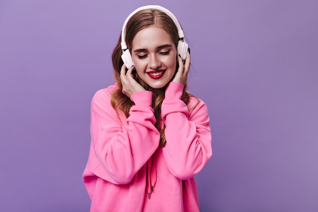 Bezpłatne zdjęcie blond dama w różowej bluzie uśmiecha się i słucha muzyki w słuchawkach