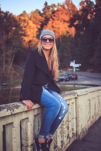 Blond kobieta w dżinsach i okularach przeciwsłonecznych siedzi na moście w parku narodowym przyrody.