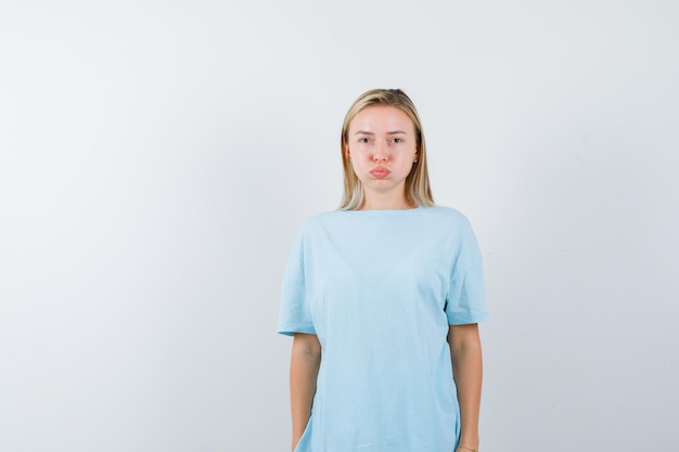 Blond kobieta w niebieskiej koszulce nadyma policzki, pozując i wyglądając na rozczarowaną