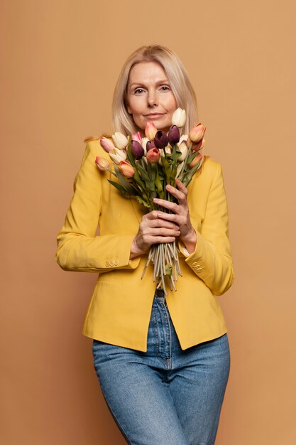 Blond starsza kobieta pozuje i trzyma kwiaty na żółtym tle