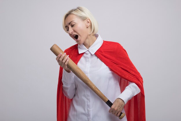 Bezpłatne zdjęcie blond superbohaterka w średnim wieku w czerwonej pelerynie trzymająca kij baseballowy, używająca go jako mikrofonu śpiewającego z zamkniętymi oczami na białym tle na białej ścianie z kopią przestrzeni