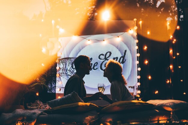 Blondynka i mąż oglądają film na dużym ekranie zawieszonym na płocie, leżąc na miejscu spoczynku z pledem i poduszkami w widoku od tyłu domku.