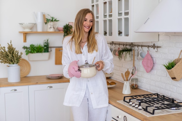 Bezpłatne zdjęcie blondynka ładna kobieta ubrana w biały lniany garnitur przygotowuje jedzenie w swojej kuchni, koncepcja idealnej żony domu.