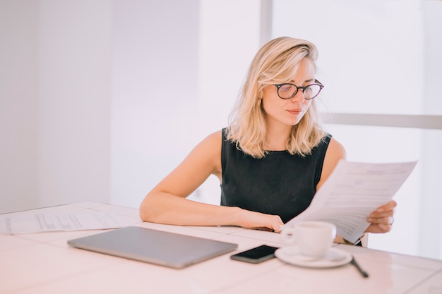 Bezpłatne zdjęcie blondynka młody bizneswoman czyta dokument przy miejscem pracy z filiżanką; laptop i telefon na stole