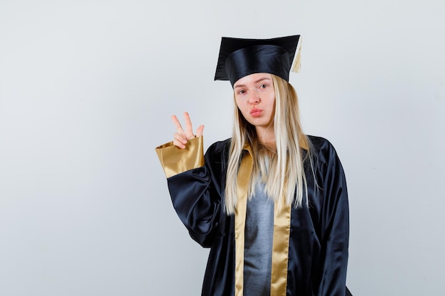 Blondynka pokazuje gest pokoju w sukni i czapce ukończenia szkoły i wygląda uroczo