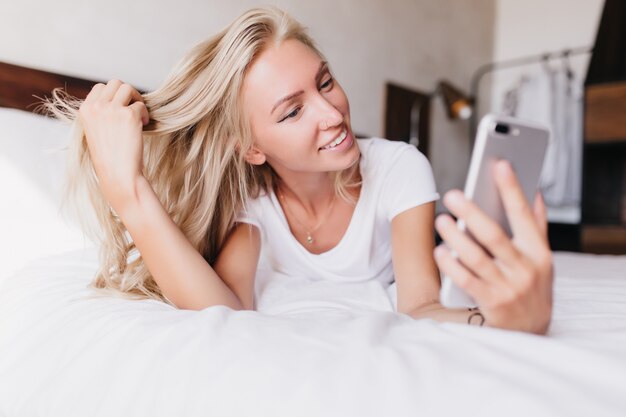 blondynka robi selfie, leżąc w łóżku. Urocza kaukaski kobieta robi sobie zdjęcie wcześnie rano.
