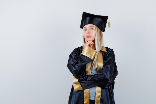 Bezpłatne zdjęcie blondynka stojąca w pozie myślenia, kładąc rękę pod brodą w sukni i czapce ukończenia szkoły i patrząc zamyślony.