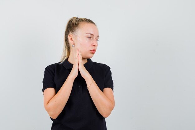 Bezpłatne zdjęcie blondynka w czarnej koszulce składając ręce i patrząc na prawą stronę i wyglądając na zmartwioną