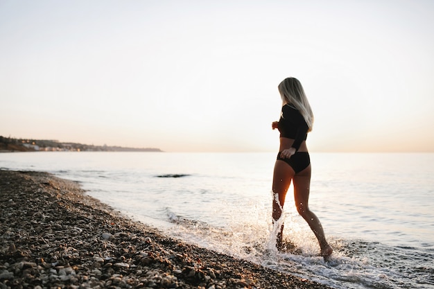 Blondynka w czarny strój kąpielowy działa na wybrzeżu oceanu na zachód słońca