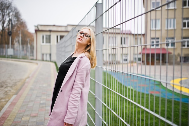 Blondynka w okularach i różowym płaszczu, czarna tunika i torebka na tle ogrodzenia na ulicy