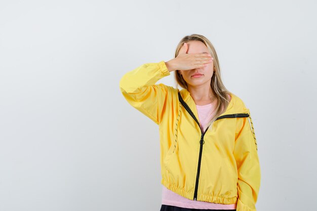 Blondynka w różowej koszulce i żółtej kurtce zakrywająca oko dłonią i wyglądająca kusząco