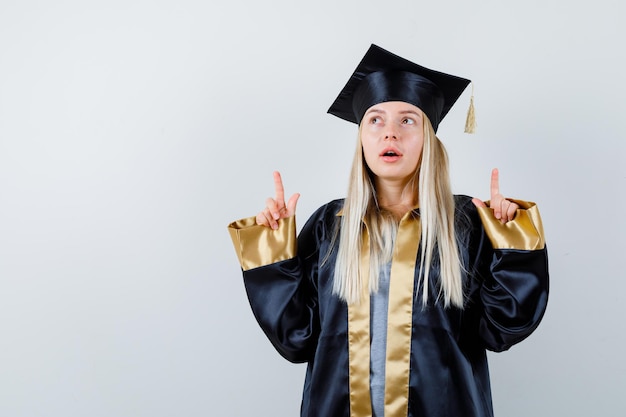 Blondynka w sukni ukończenia szkoły i czapce skierowana w górę palcami wskazującymi i wyglądająca uroczo