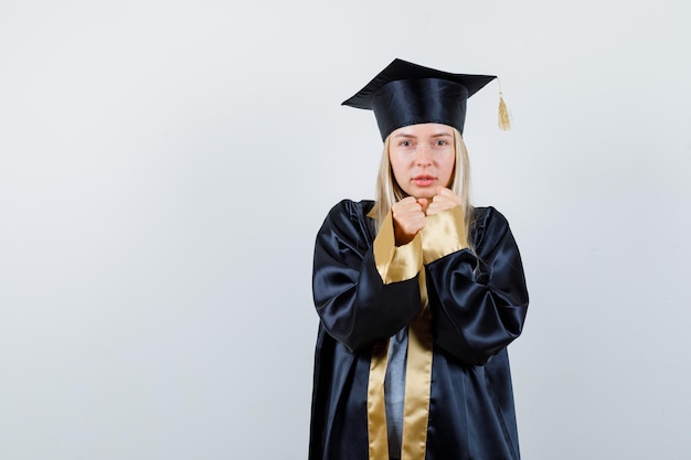 Blondynka w sukni ukończenia szkoły i czapce stoi w pozie bokserki i wygląda na potężną