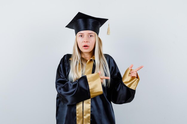 Blondynka w sukni ukończenia szkoły i czapce wskazująca palcem wskazującym w prawo i wyglądająca na zaskoczoną