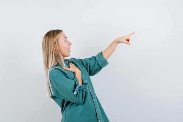 Bezpłatne zdjęcie blondynka w zielonej bluzce trzymająca rękę na klatce piersiowej, wskazująca w prawo i wyglądająca na zaskoczoną