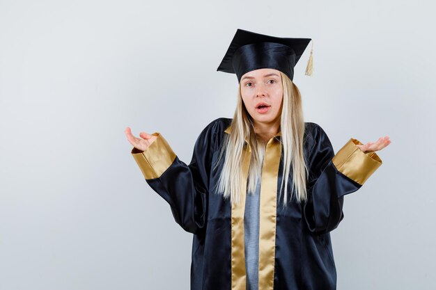 Blondynka wyciągając ręce w sposób przesłuchania w sukni i czapce ukończenia szkoły i patrząc zbita z tropu.