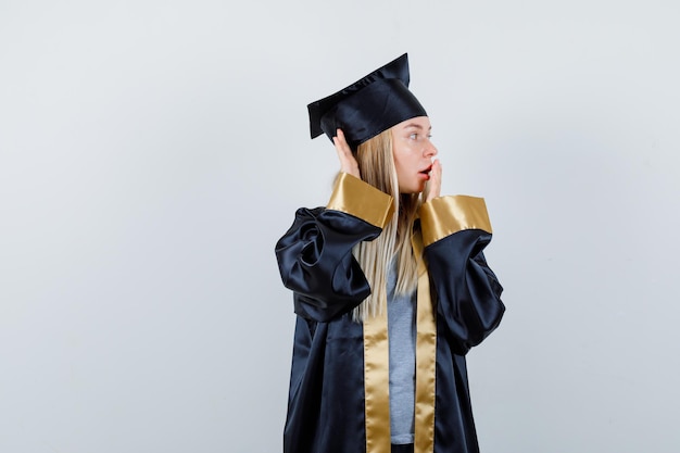 Blondynka zakrywająca usta dłonią, trzymająca rękę przy uchu, aby usłyszeć coś w sukni ukończenia szkoły