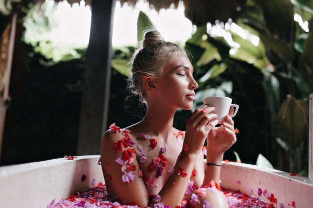 Blondynka zrelaksowany kobieta pije kawę siedząc w wannie.