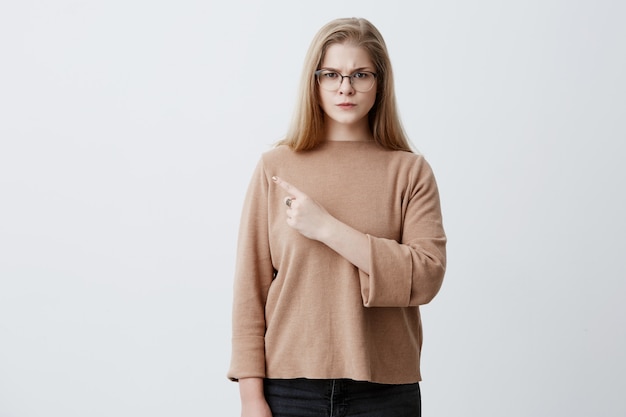 Blondynki kobieta wskazuje z palcem przy pustą ścianą z kopii przestrzenią dla reklamy teksta lub produktu w brown pulowerze i eyeglasses, patrzeje kamerę z poważnym wyrażeniem. Koncepcja reklamy