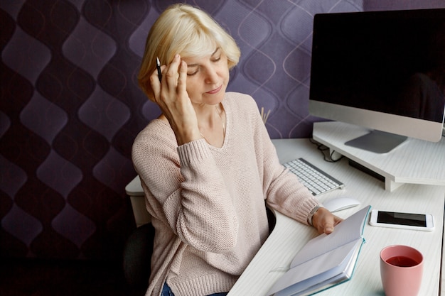 Bezpłatne zdjęcie blondyny dorośleć kobiety writing w jej planistyce podczas gdy pracujący komputerem w domu