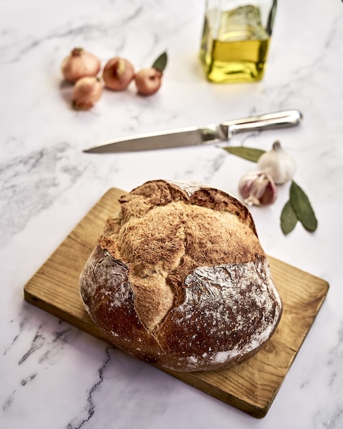 Bochenek świeżo upieczonego ciemnego chleba na drewnianej desce z cebulą, czosnkiem, oliwą i nożem