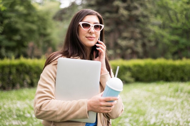 Boczny widok kobieta trzyma laptop i napój outdoors z okularami przeciwsłonecznymi