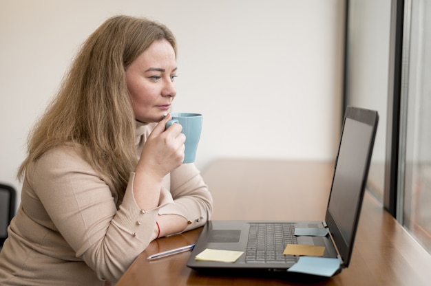 Boczny widok kobiety mienia kubek i działanie na laptopie przy biurem