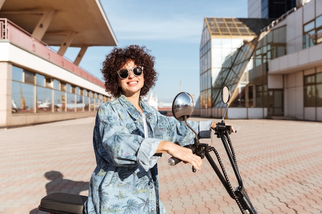 Boczny widok uśmiechnięta kędzierzawa kobieta w okularach przeciwsłonecznych pozuje na nowożytnym motocyklu outdoors