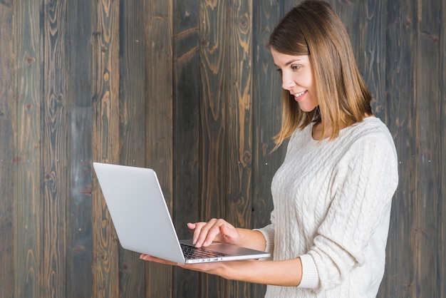 Boczny widok uśmiechnięta młoda kobieta używa laptop przeciw drewnianej ścianie
