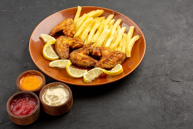 Boczny widok z bliska talerz fastfood apetyczne frytki skrzydełka z kurczaka i cytryna z trzema rodzajami sosów na ciemnym tle