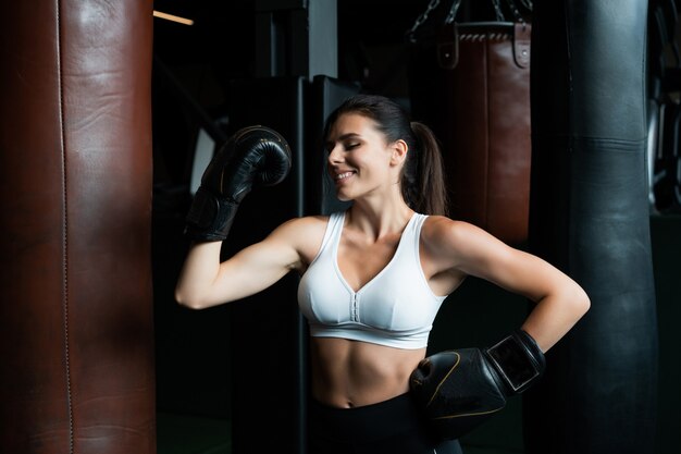 Bokserska kobieta pozuje z worek treningowy, na ciemnym gym. Silna i niezależna koncepcja kobiety