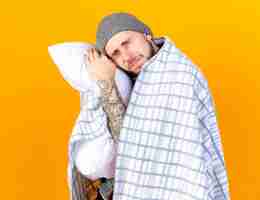Bezpłatne zdjęcie bolący młody chory ubrany w zimową czapkę i szalik owinięty w kratę trzyma i kładzie głowę na poduszce na pomarańczowej ścianie
