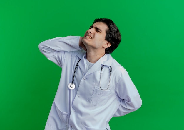Bolący młody mężczyzna lekarz ubrany w medyczny szlafrok i stetoskop, trzymając ręce na szyi i plecach na białym tle na zielonej ścianie z miejsca na kopię