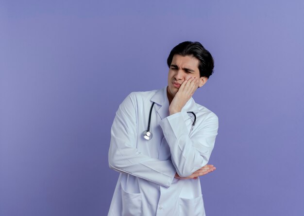 Bolący młody mężczyzna lekarz ubrany w szlafrok i stetoskop trzymając rękę na policzku z zamkniętymi oczami cierpiących na ból zęba na białym tle