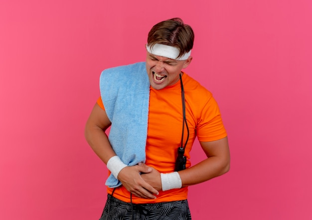 Bezpłatne zdjęcie bolący młody przystojny sportowy mężczyzna w opasce i opaskach na nadgarstek z ręcznikiem i skakanką wokół szyi, trzymając brzuch z zamkniętymi oczami cierpiącego na ból odizolowany na różowo z miejscem na kopię